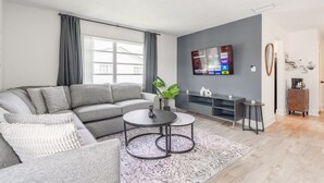 Uma sala de estar moderna com um sofá seccional cinza, duas mesas redondas, uma TV de tela plana na parede e um pequeno aparador.