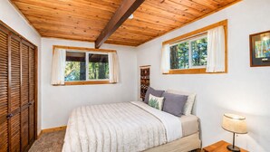 Ein Schlafzimmer mit Holzboden, zwei mit Vorhängen versehene Fenster, ein Bett mit flauschiger Decke, ein Nachttisch mit Lampe und ein Holzschrank.