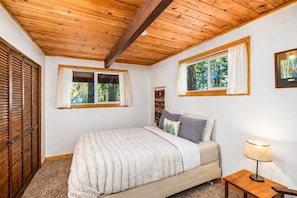 A bedroom with a wooden ceiling, two windows with curtains, a bed with a fluffy blanket, a bedside table with a lamp, and a wooden wardrobe.