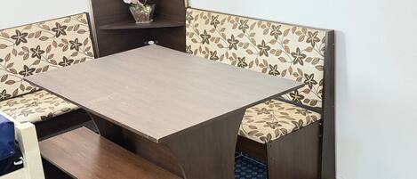 A corner dining table with floral-patterned cushioned benches, a small potted orchid, and a wooden shelf above.