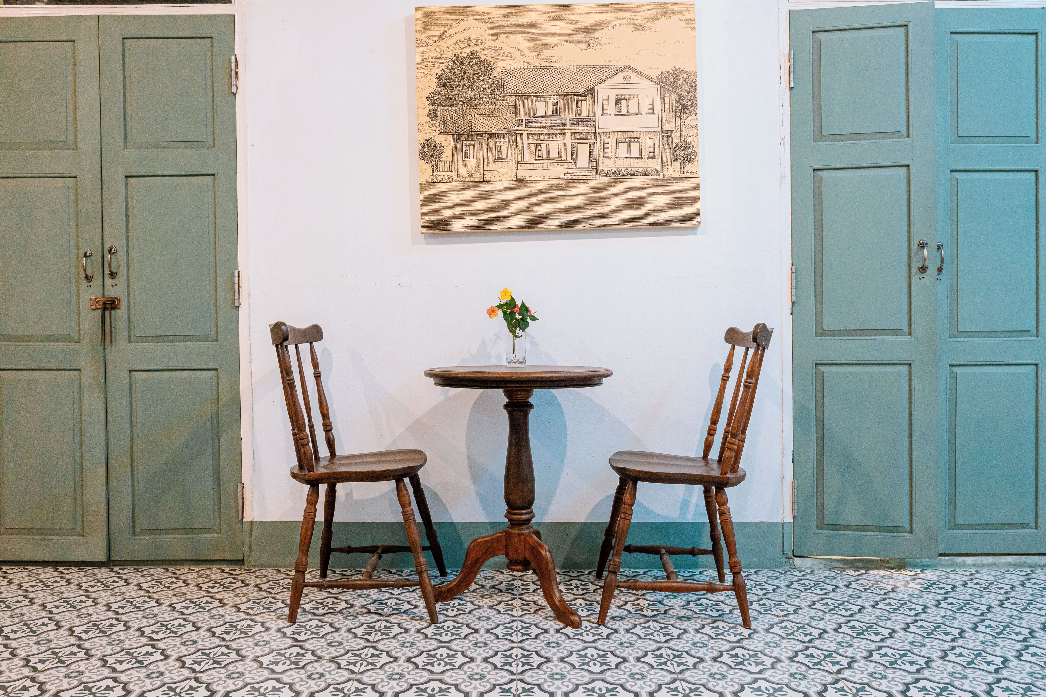 A room with two wooden chairs around a small round table, a painting on the wall, and two doors.