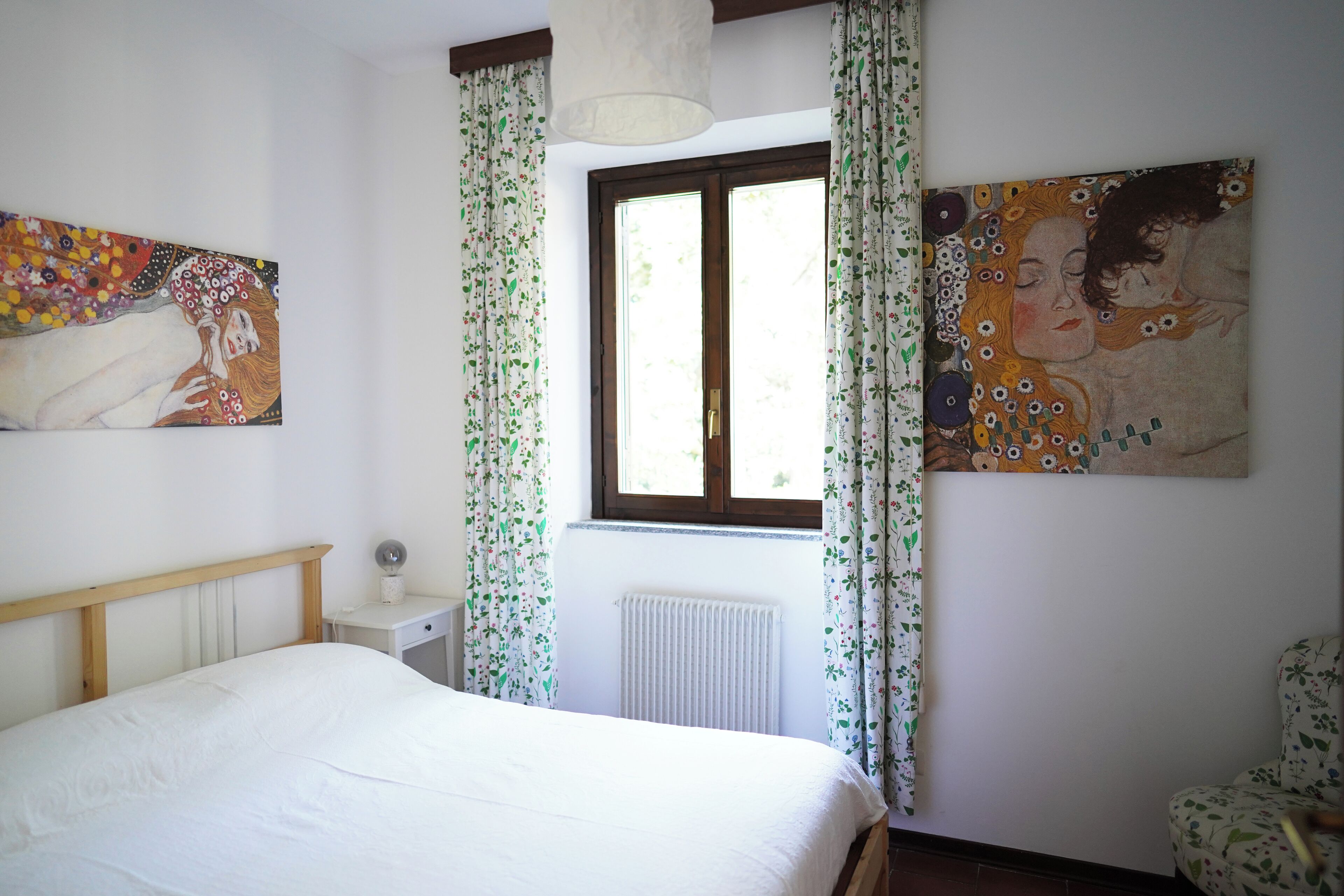 Ein Schlafzimmer mit einem Bett, einem Nachttisch, einer Lampe, einem Fenster mit Vorhängen und zwei Bildern an der Wand.