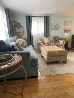 A living room with a sofa, an ottoman, a round table with a plant, and a bookshelf with various items.