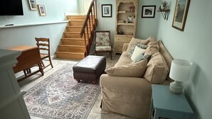 A living room with a sofa, ottoman, and a small table with a lamp. There is a staircase leading to another level.
