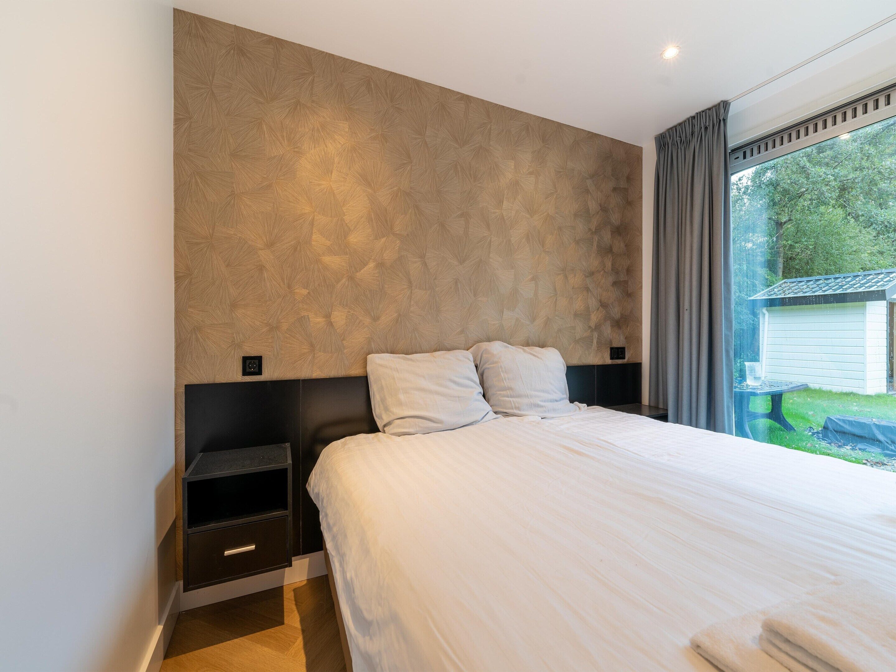 A modern bedroom with a large bed, a headboard with a geometric pattern, a nightstand, and a view of a house through a window.