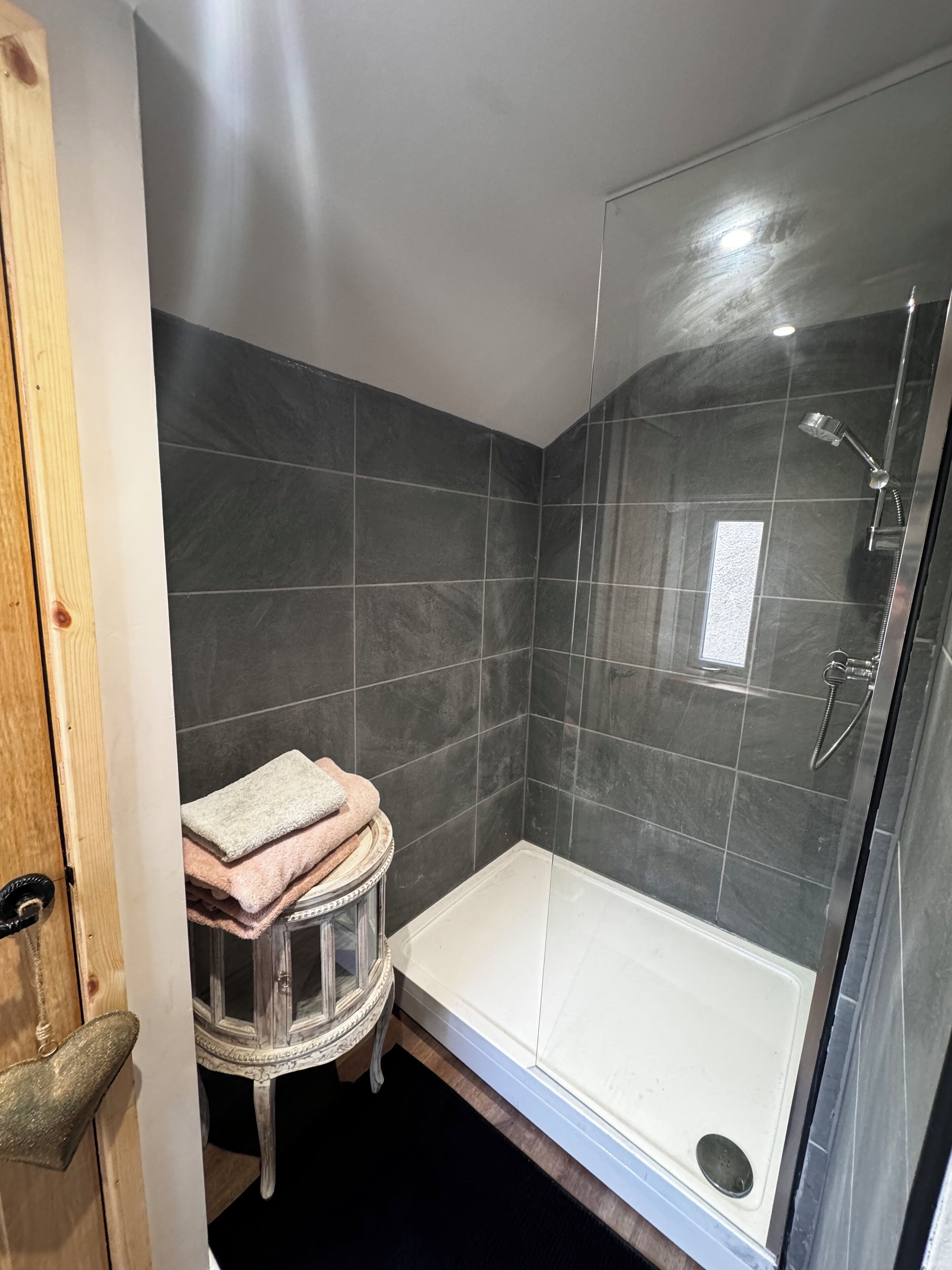 A modern bathroom with dark tiles, a glass shower enclosure, and a small round stool with towels.