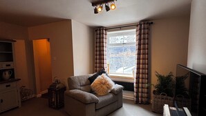 Una sala de estar acogedora con un sofá, un tapete pequeño, una mesa de centro de madera, un televisor y una ventana con cortinas.