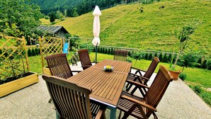Un conjunto de mesa y sillas de madera en un patio con vistas a una ladera verde y una cabaña de madera al fondo.