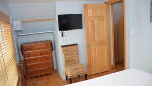 Ein Schlafzimmer mit einem Holzkasten, einem Korbstuhl, einem Bett, einem an der Wand befestigten Fernseher und einer Holztür.