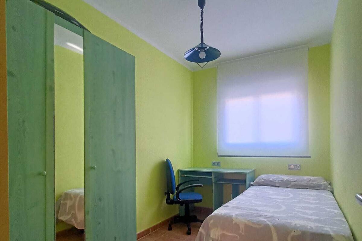 A room with a bed, a desk, and a chair. There is a window with blinds and a ceiling fan.