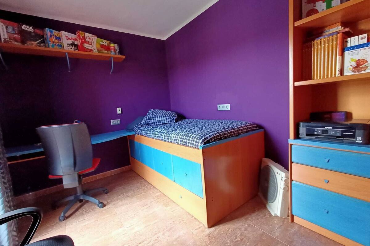 A compact bedroom with a bed, desk, chair, and shelves with books and a printer.