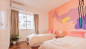 Une chambre d’hôtel avec deux lits, une plante en pot, une fenêtre avec des rideaux et une fresque murale colorée.