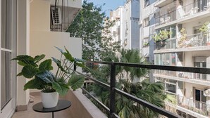 Un balcon agrémenté d’un pot de fleurs, d’une petite table et offrant une vue sur les bâtiments et la végétation environnants.