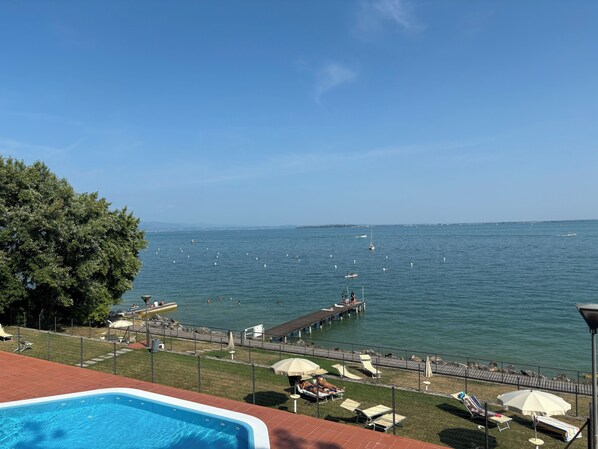 A pool area with lounge chairs and umbrellas, a dock extending into the water, and a clear view of the sea and distant shoreline.