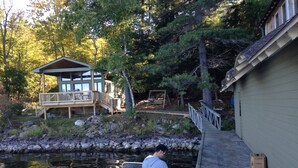 A wooden dock leading to a small boat, a cabin with a deck, and a rocky shoreline.
