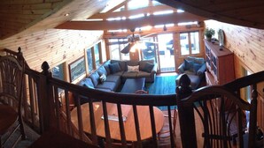 A wooden cabin interior with a loft bedroom, a dining area, and a living room with a fireplace.