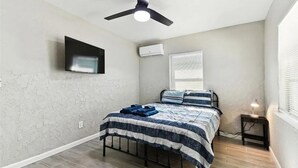 Un dormitorio con cama, ventilador de techo, televisor montado, aire acondicionado, ventana con persianas y lámpara de noche.