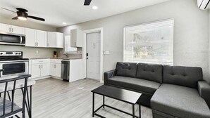 Una cocina moderna con armarios blancos, microondas y estufa. En la zona de estar, hay un sofá gris y una mesita.