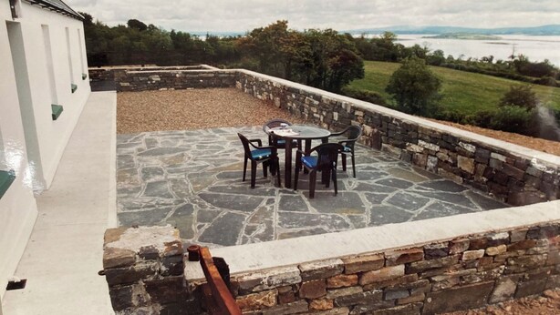 Un patio de piedra con mesa y sillas, rodeado por un muro de piedra, con vistas a un paisaje pintoresco.