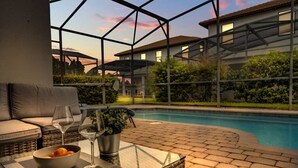 Zona junto a la piscina con mesa de vidrio, mobiliario de mimbre y vistas a un edificio y follaje.
