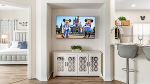 Una sala de estar con un televisor montado sobre un mueble blanco, un comedor con un taburete de bar, y una cama con vistas a la habitación.