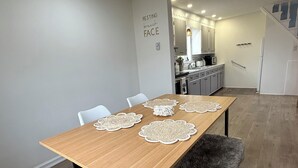 A dining area with a wooden table, white chairs, and a fur throw. There is a kitchen in the background with white cabinets and a staircase.