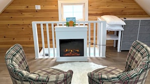 An attic bedroom with a fireplace, plaid chairs, a white rug, and a wooden ceiling.