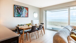 A modern living room with a dining area, a large abstract painting, and a balcony with a beach view.