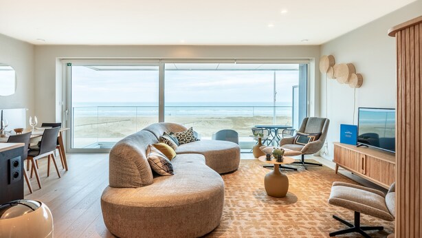 A spacious living area with a large window overlooking a beach, a cozy seating arrangement, and a television on a wooden cabinet.