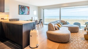 A modern living room with a large window overlooking a beach, a round coffee table, and a sofa with cushions.