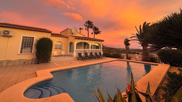 A pool area with a house in the background, surrounded by palm trees and a sunset sky. - Villa Casa Creativa: your sunny dream holiday home with breathtaking sea views (El Poble Nou de Benitatxell)