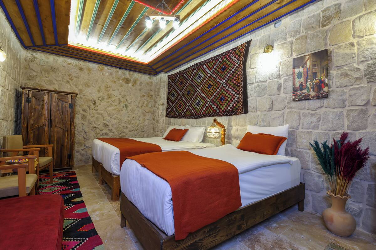 A room with two beds, a wooden wardrobe, a chair, a patterned rug, and a stone wall.
