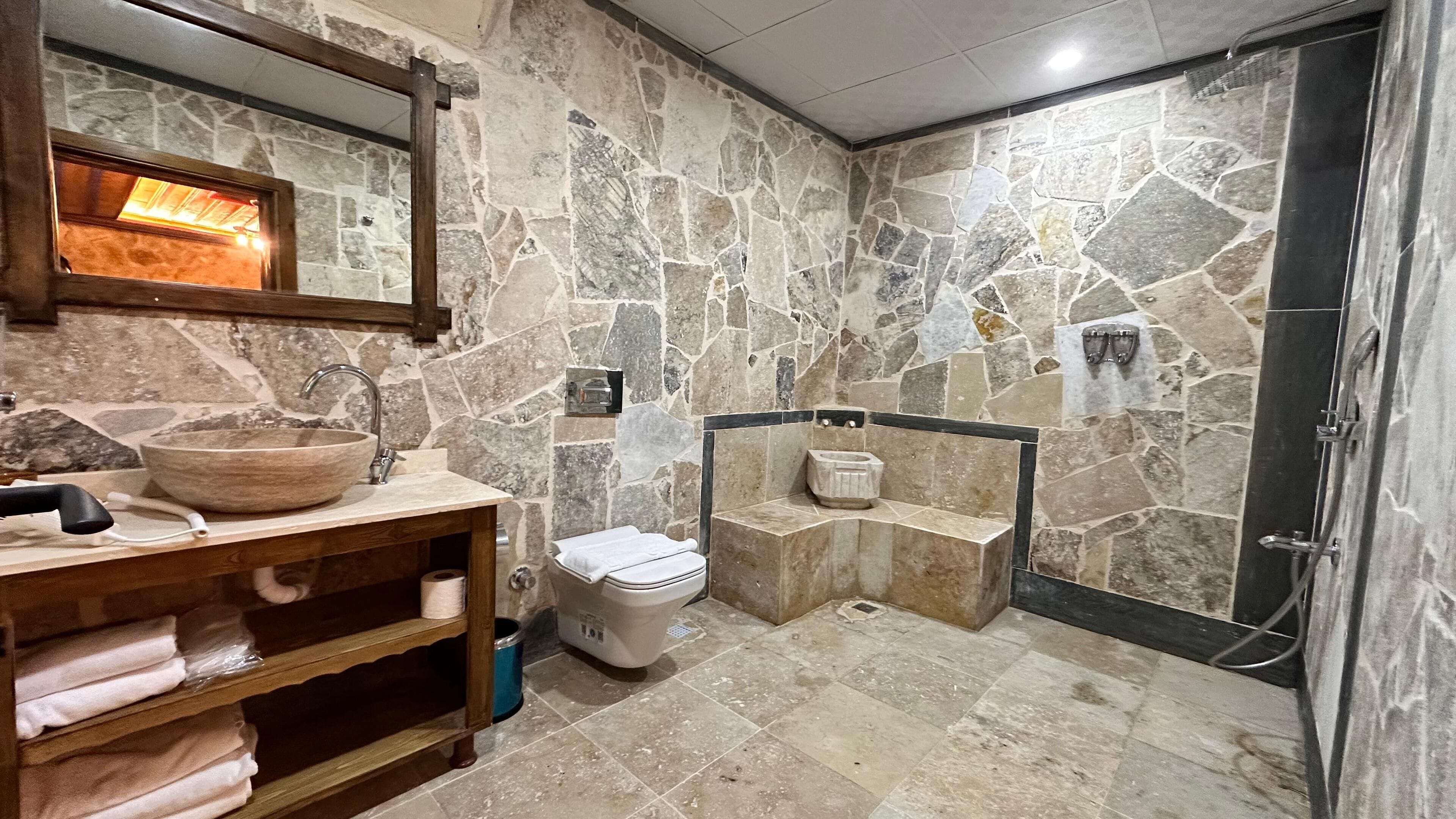 Un baño con pared de piedra, tocador de madera, un lavabo grande, inodoro y zona de ducha.
