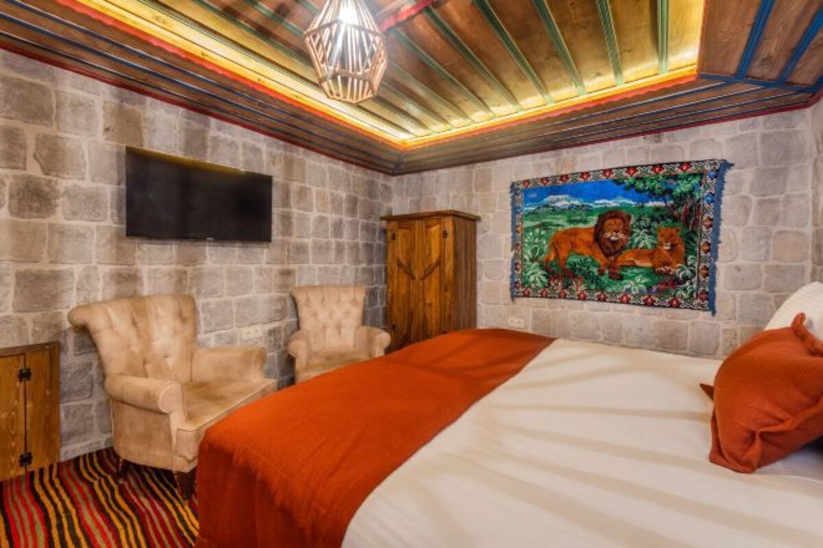 A bedroom with a stone wall, a bed with an orange blanket, a wooden cabinet, a beige armchair, and a painting of lions.