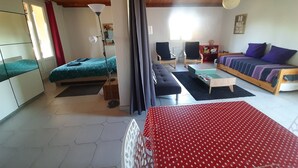 A room with a bed, a sofa, a desk, and a chair. There is a window with curtains, a lamp, and a wall clock.