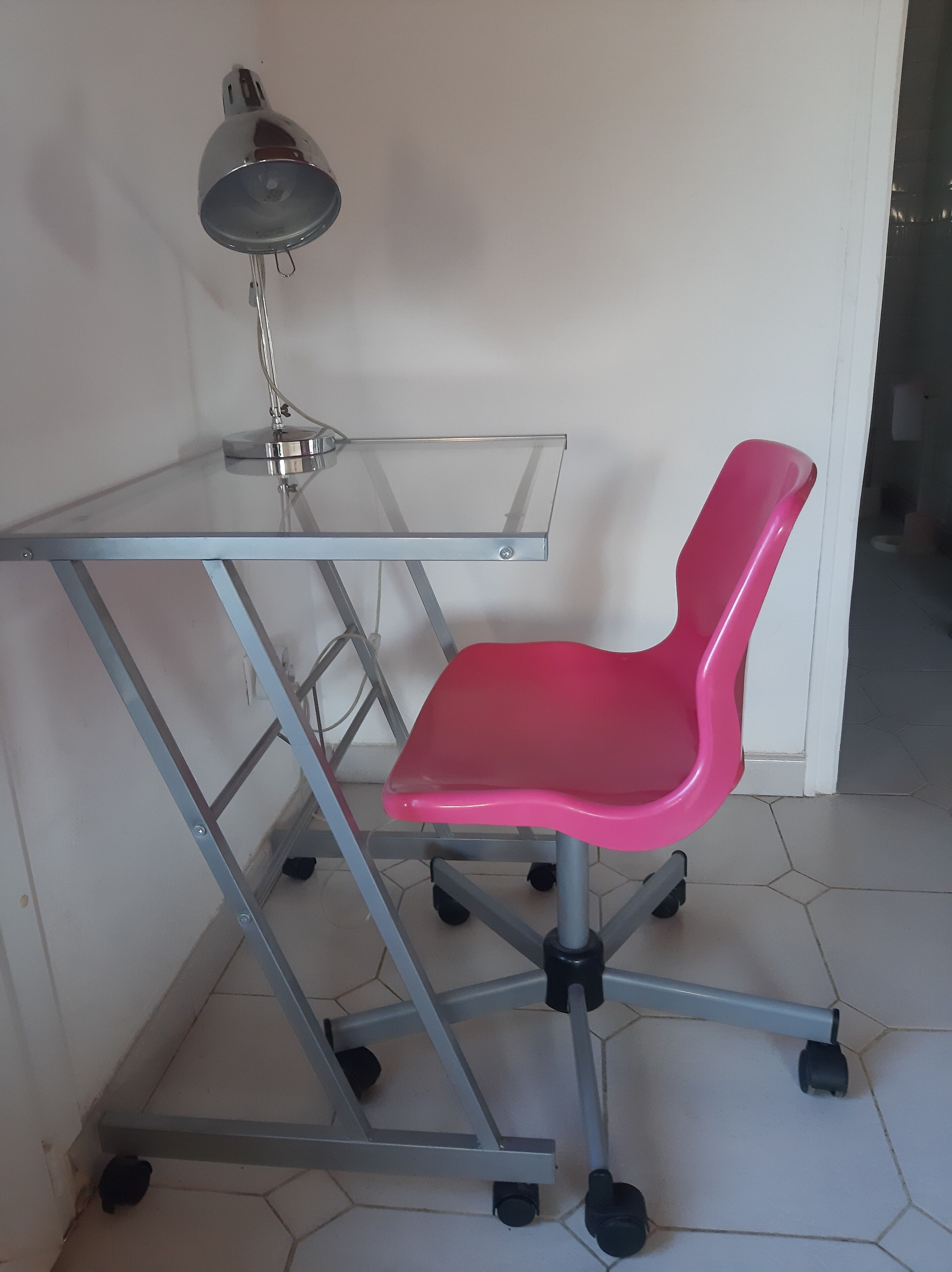 Un bureau avec un plateau en verre transparent, une chaise rose à roulettes et une lampe de bureau.