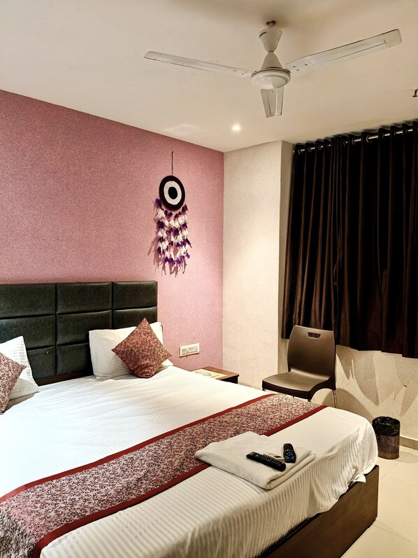 A hotel room with a large bed, a ceiling fan, a wall-mounted dreamcatcher, a chair, and a window with curtains. - HOTEL SUNRISE CAVE (Surat)