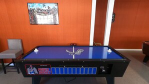 Un biliardo con superficie in feltro blu, una biglia bianca e un logo. A sinistra, una sedia e al muro, un quadro.