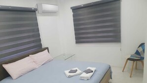 Un dormitorio con cama, almohadas, una silla y un ventanal con persianas.