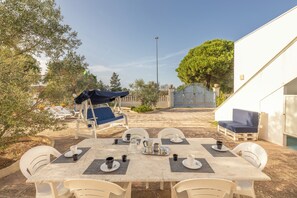 An outdoor dining area with a table set for four, surrounded by white chairs, and a blue umbrella providing shade. - Beautiful villa surrounded by greenery   (Torre Suda)