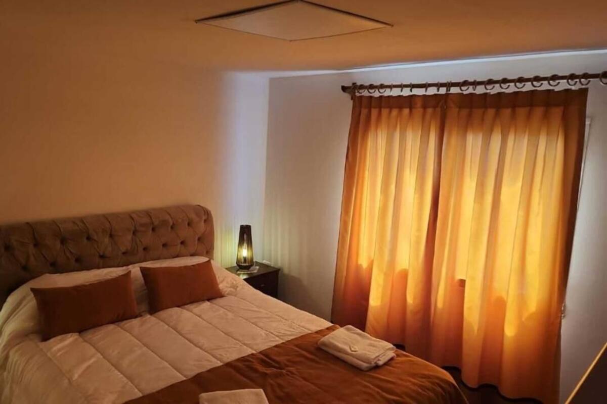 Una cama bien hecha con cabecera, lámpara de noche y ventana con cortinas.