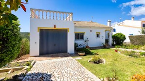 A white building with a tiled roof, a stone-paved entrance, and a garden with various plants.