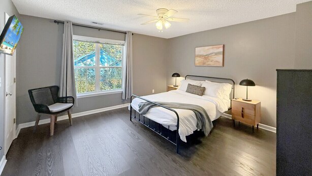 Ein Schlafzimmer mit Bett, Sessel, Nachttisch, Deckenventilator und einem Fenster mit Vorhängen.