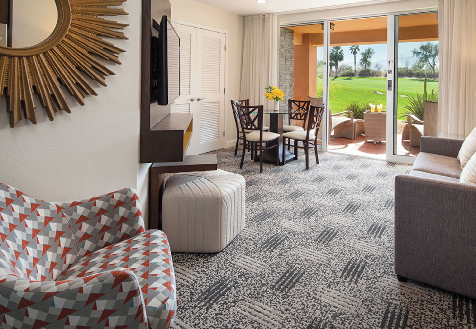 A living room with a patterned carpet, a sofa, a coffee table with chairs, and a view of a golf course through a sliding glass door.