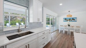 A modern kitchen with white cabinetry, a large window, and a dining area with a table and chairs.