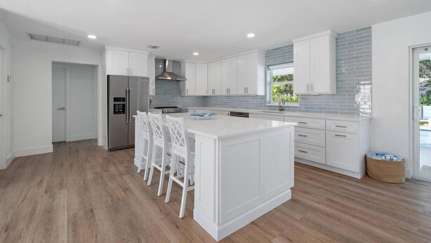 A modern kitchen with white cabinetry, a central island with bar stools, a stainless steel refrigerator, and a window with a view of greenery.