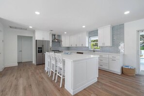 A modern kitchen with white cabinetry, a central island with bar stools, a stainless steel refrigerator, and a window with a view of greenery.