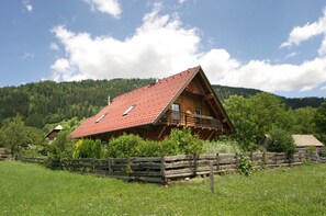 A wooden cabin with a red roof, surrounded by a wooden fence and lush greenery.