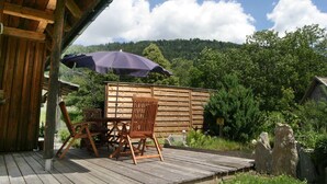 Eine Holzterrasse mit Tisch und Stühlen, einem violetten Sonnenschirm und Blick auf einen bewaldeten Hügel.