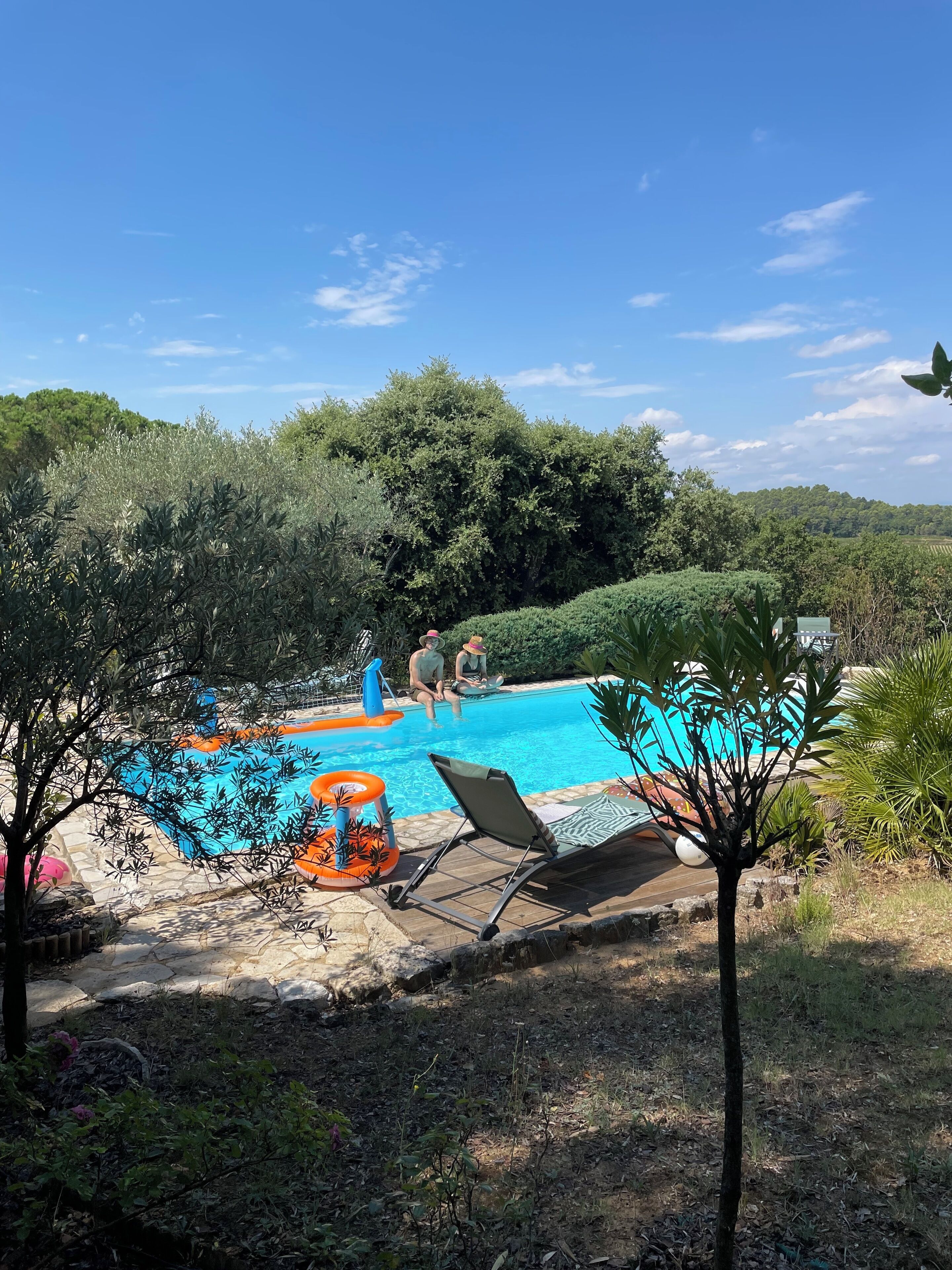 Une piscine entourée de verdure, avec quelques chaises longues et une personne assise au bord de la piscine.